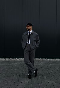 Γκρίζα ζακέτα με φερμουάρ πάνω από άσπρο πουκάμισο και μαύρη γραβάτα, συνδυασμένη με γκρίζα παντελόνια και μαύρα παπούτσια. Ο φόντος περιλαμβάνει ένα σκοτεινό τοίχο και pave.