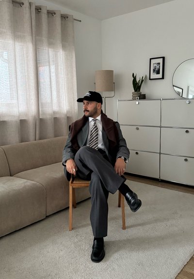 Traje de negocios gris con chal marrón, camisa blanca y corbata a rayas. Gorra negra y zapatos negros. Sentado en una silla de madera, sofá beige cerca.