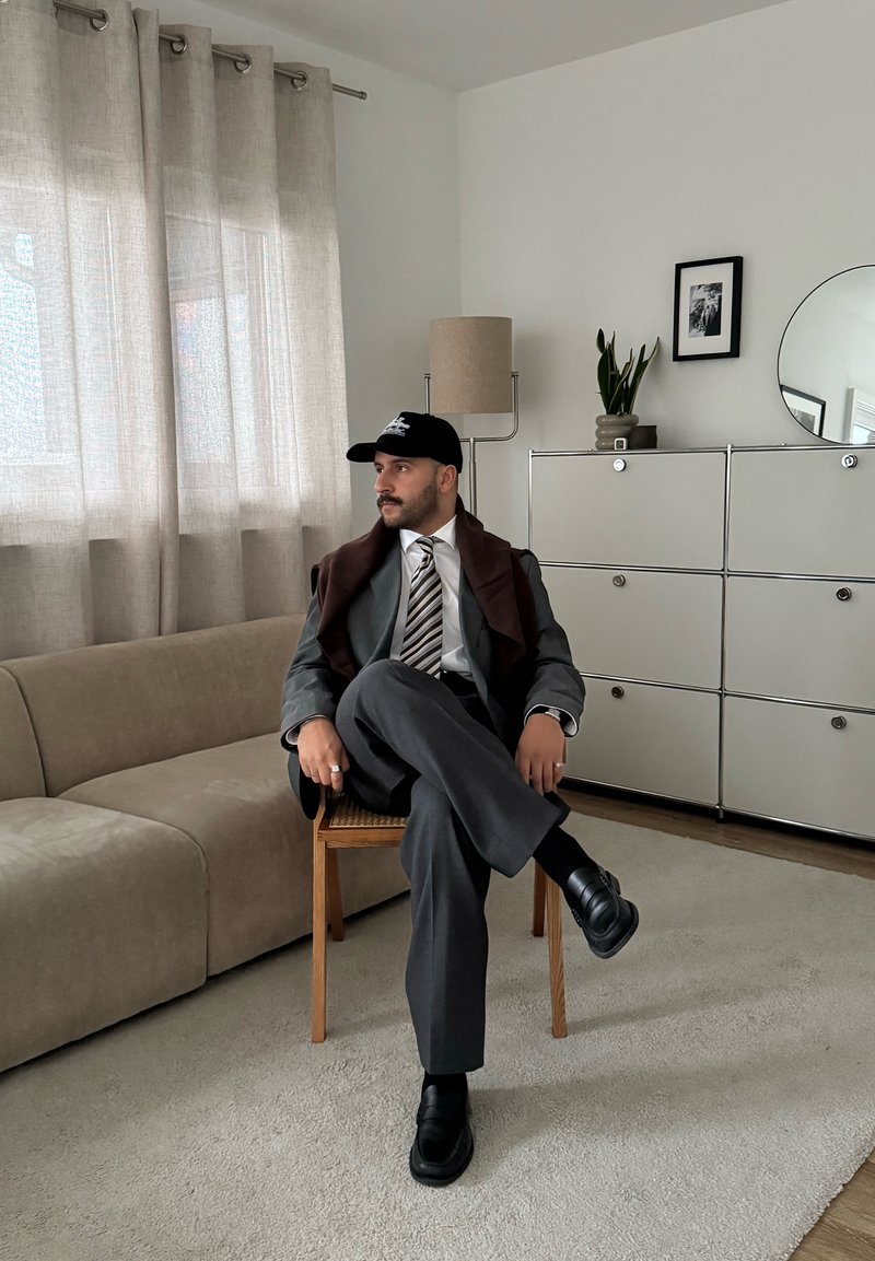 Costume d'affaires gris avec un châle marron, chemise blanche et cravate rayée. Casquette noire et chaussures noires. Assis sur une chaise en bois, canapé beige à proximité.