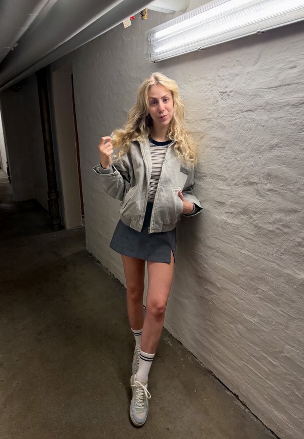 Tânără cu păr blond lung, purtând o jachetă gri, top în dungi, fustă bleumarin, șosete albe și adidași, stând într-un hol întunecat.