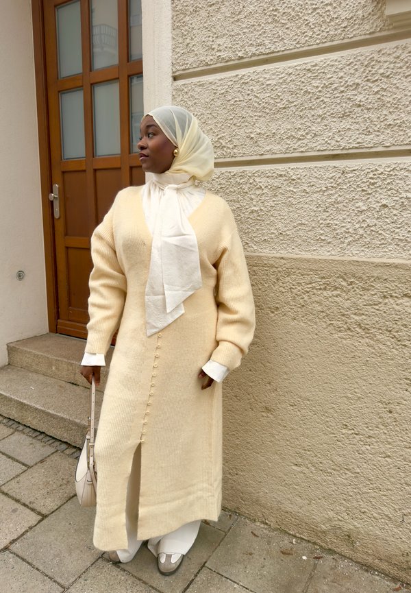 Vrouw draagt een beige lange cardigan, een witte hoofddoek, oorbellen en houdt een witte handtas vast, staand bij een getextureerde beige muur naast een houten deur.