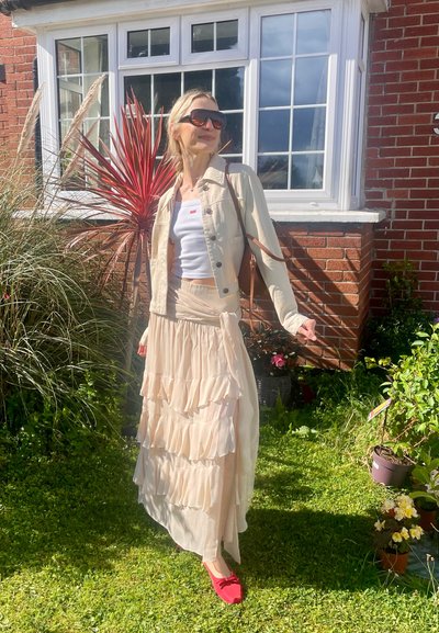 Mujer con gafas de sol, chaqueta ligera, camiseta blanca y falda beige fluida, de pie sobre el césped junto a una casa de ladrillo y plantas de jardín.