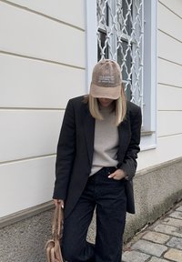 Άτομο με ξανθά μαλλιά που φοράει μπεζ καπέλο, γκρι πουλόβερ, μαύρο σακάκι και σκούρο τζιν στέκεται δίπλα σε έναν λευκό τοίχο κρατώντας μια μπεζ τσάντα.