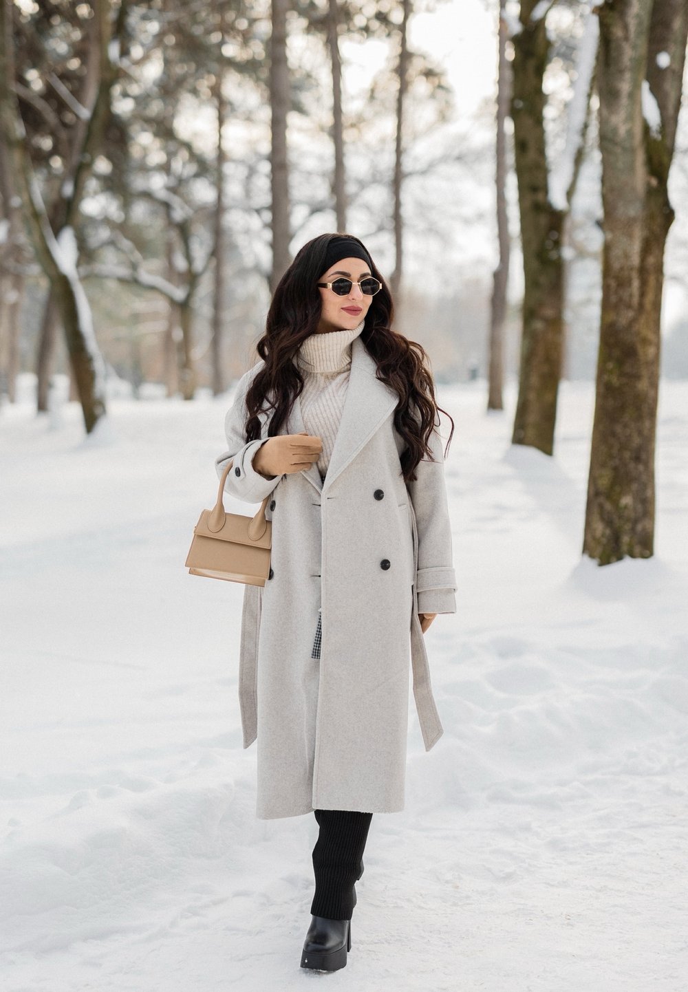 Femme portant un long manteau gris clair, des bottes noires et des lunettes de soleil, marchant dans une forêt enneigée en portant un sac à main beige.