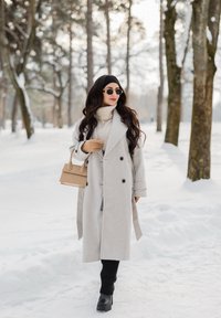 Femme portant un long manteau gris clair, des bottes noires et des lunettes de soleil, marchant dans une forêt enneigée en portant un sac à main beige.
