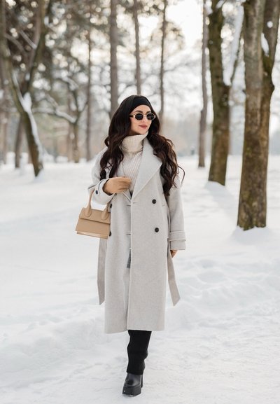 Mujer con un abrigo largo gris claro, botas negras y gafas de sol caminando por un bosque nevado, llevando un bolso beige.