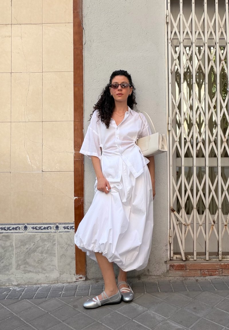 Femme aux cheveux bouclés portant des lunettes de soleil, une robe blanche, des chaussures argentées et un sac à main beige, se tient contre un mur et une grille métallique sur un trottoir carrelé.