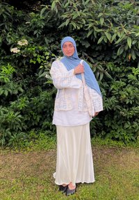 Hvit langermet bluse, lys blå mønstrete cardigan, lang kremfarget skjørt og blått hodesjal. En vevd veske henger fra skulderen.
