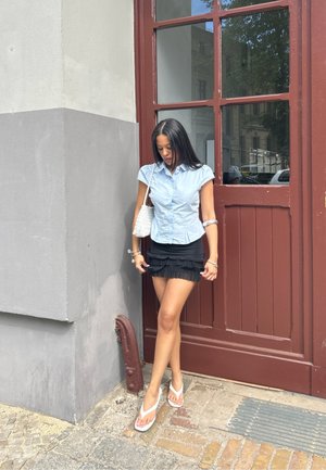 Camisa de manga corta azul claro, mini falda negra con volantes, chanclas blancas y bolso de mano blanco de fibra trenzada. De pie cerca de una puerta roja.