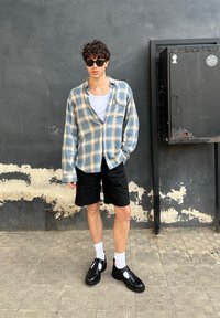 Camicia a scacchi blu e grigia con bottoni, pantaloni corti neri, scarpe nere con calzini bianchi. Sfondo semplice con una scatola sulla parete.