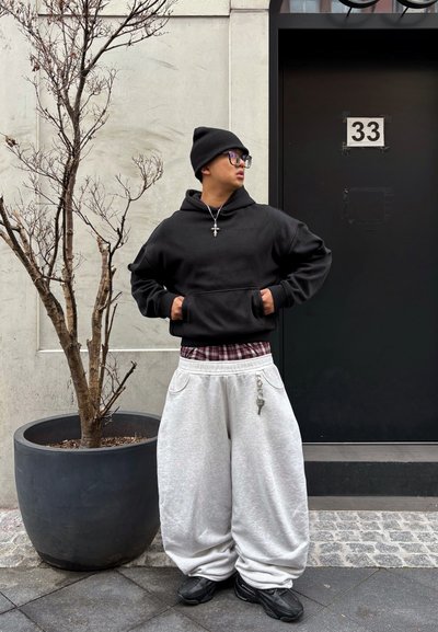 Joven con gorro negro, gafas, sudadera negra con capucha, pantalones grises anchos y zapatos negros, de pie junto a un gran árbol sin hojas en maceta junto a una puerta negra.
