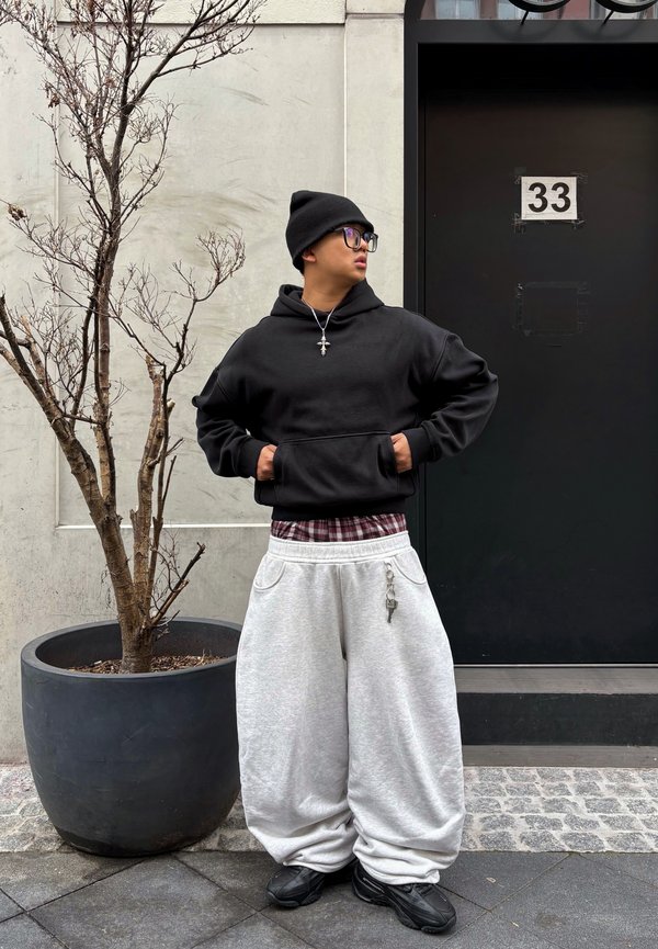 Junge Person, die eine schwarze Mütze, Brille, schwarzen Hoodie, weite graue Hose und schwarze Schuhe trägt, steht neben einem großen, entlaubten Baum in einem Topf neben einer schwarzen Tür.