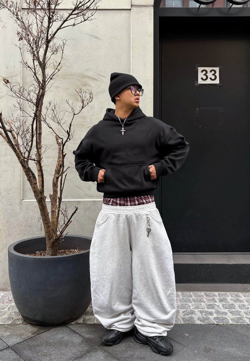 O persoană tânără purtând căciulă neagră, ochelari, hanorac negru, pantaloni gri largi și pantofi negri stă lângă un copac mare fără frunze într-un ghiveci, lângă o ușă neagră.