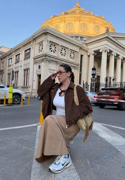 Mujer con gafas, chaqueta marrón, falda beige y zapatillas sentada en un bordillo con un edificio histórico con columnas y una cúpula dorada al fondo.