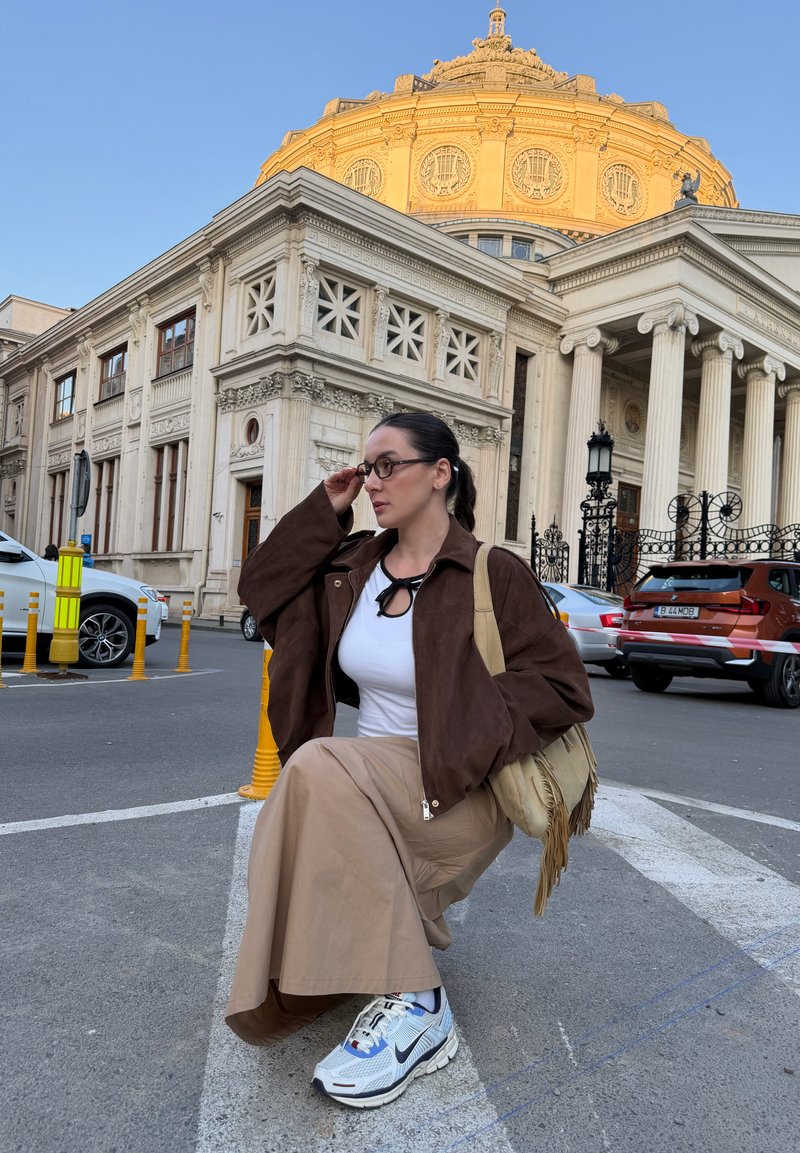 Femme portant des lunettes, une veste marron, une jupe beige et des baskets assise sur un trottoir avec un bâtiment historique avec des colonnes et un dôme doré en arrière-plan.