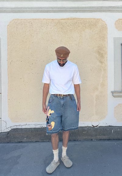 Pantalones cortos de denim de color azul deslavado, con un gran diseño abstracto multicolor en la pierna izquierda. Combinados con una camiseta blanca y una boina color beige.