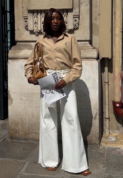 Camisa beige de botones, pantalones de mezclilla anchos blancos, bolso marrón y cartel impreso. Usado a la luz del sol contra una pared texturizada.