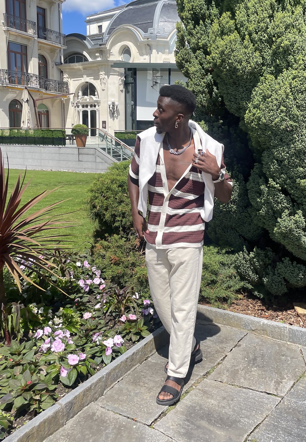 Μακρυμάνικη μπλούζα από ύφασμα σε μπορντό και λευκές ρίγες, μπεζ παντελόνι, μαύρα σανδάλια. Στέκεται σε έναν κήπο με πράσινα φυτά και ροζ λουλούδια.