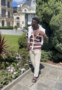 Μακρυμάνικη μπλούζα από ύφασμα σε μπορντό και λευκές ρίγες, μπεζ παντελόνι, μαύρα σανδάλια. Στέκεται σε έναν κήπο με πράσινα φυτά και ροζ λουλούδια.