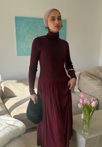 Femme portant une robe à col roulé à manches longues marron rougeâtre et un foulard beige, tenant un chapeau noir dans un salon avec des tulipes roses sur la table.