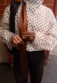 Persona vistiendo una blusa blanca con lunares y una corbata marrón, sosteniendo unas gafas de sol de carey con lentes ámbar y con una correa de mochila marrón visible.