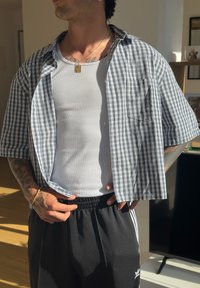 Camisa de manga curta, xadrez em azul e branco, usada aberta sobre uma camisola justa cinza-claro. Calças pretas com cintura elástica.