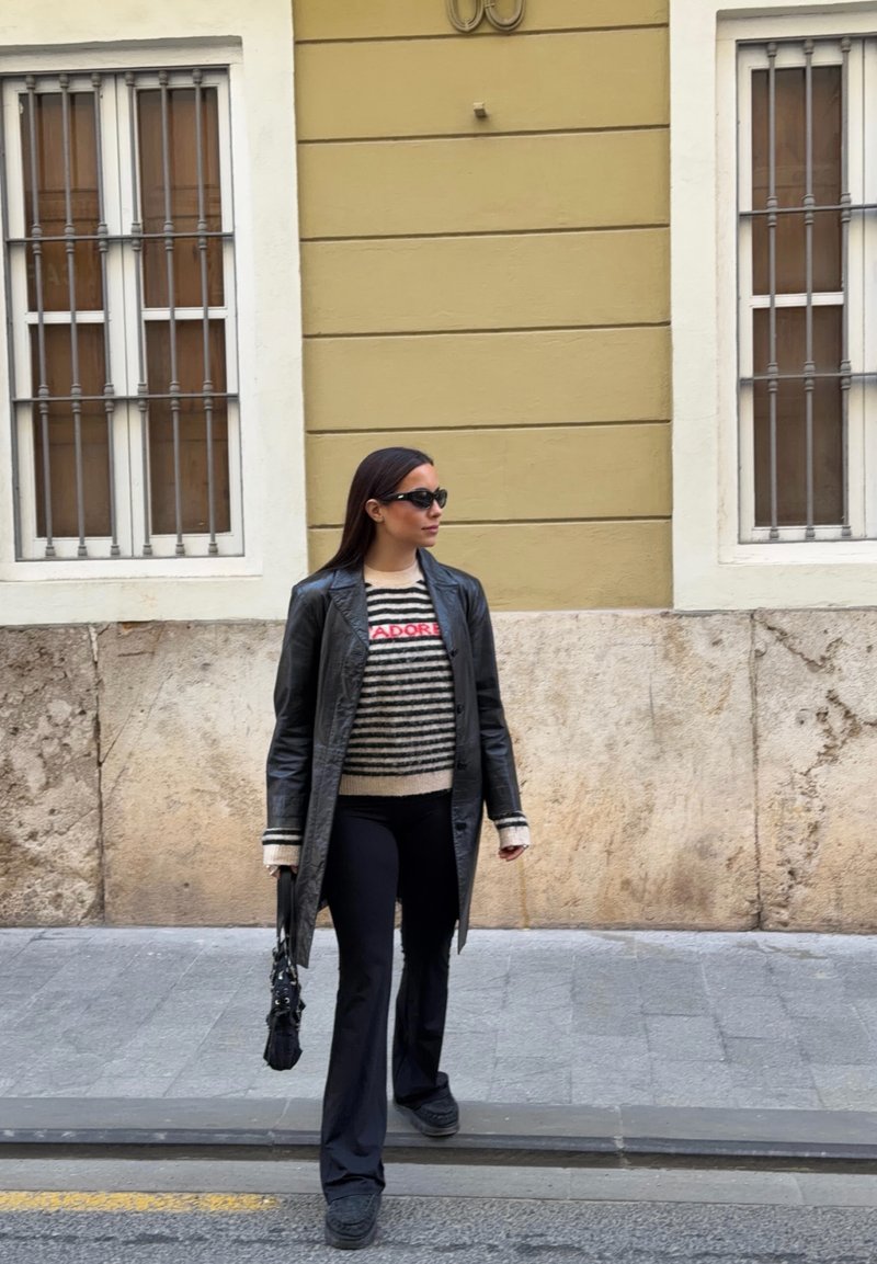 Femme portant des lunettes de soleil, un manteau en cuir noir, un pull rayé avec le texte « J'ADORE » et un pantalon noir, marchant sur un trottoir en ville près de fenêtres grillagées.
