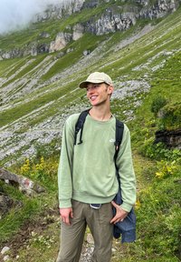 Mladý muž v svetle zelené mikině a čepici se usmívá při pěší túře po kamenitém, travnatém horském chodníku s útesy pokrytými mlhou.