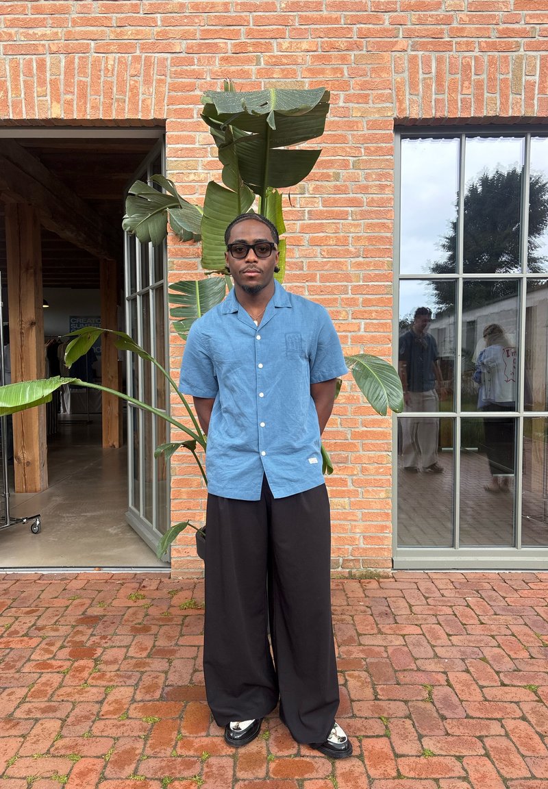 Homme en chemise bleue à manches courtes et pantalon large noir se tient devant un mur en briques avec de grandes feuilles vertes et des fenêtres en verre reflétant deux personnes.