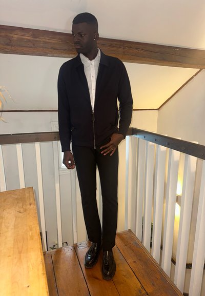 Hombre con chaqueta negra, camisa blanca, pantalones negros y zapatos negros de pie sobre un suelo de madera cerca de una barandilla blanca en el interior.