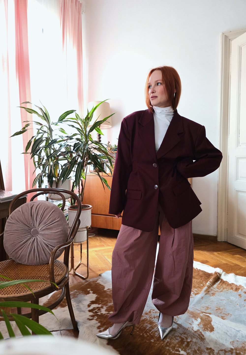 Frau mit rotem Haar, die einen oversized dunklen Blazer, einen weißen Rollkragenpullover, weite mauvefarbene Hosen und silberne Absätze trägt, steht in einem hellen Raum mit Pflanzen und Holzmöbeln.