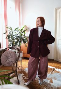 Frau mit rotem Haar, die einen oversized dunklen Blazer, einen weißen Rollkragenpullover, weite mauvefarbene Hosen und silberne Absätze trägt, steht in einem hellen Raum mit Pflanzen und Holzmöbeln.