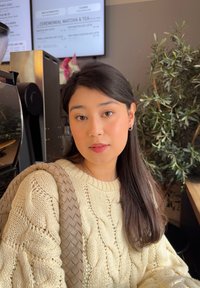 Maglione a trecce color crema abbinato a una borsa beige intrecciata. La luce naturale esalta i capelli lisci e i tratti del viso delicati. Sullo sfondo si vedono delle piante e un menù.