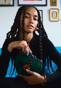 Femme aux longues tresses torsadées portant des boucles d'oreilles rouges et un haut noir, tenant une pochette verte brodée d'une planète colorée, assise sur un canapé bleu.