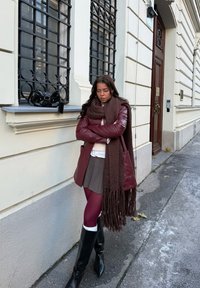 Veste en cuir marron, écharpe à franges marron, pull blanc, jupe grise, collants bordeaux et bottes noires montantes jusqu'aux genoux. Sac noir posé sur le rebord de la fenêtre.