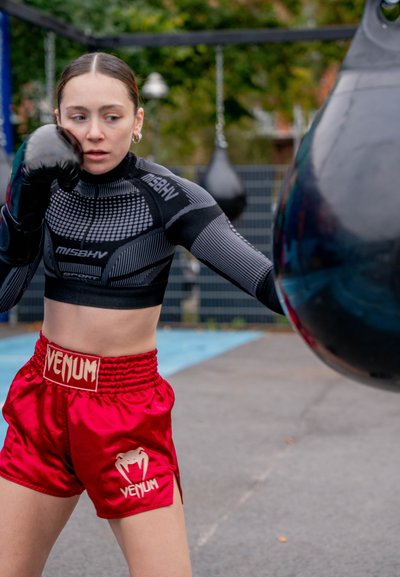 Boxeador con una camiseta ajustada negra con detalles de malla y pantalones cortos de satén rojos, con el logo "VENUM", golpeando un saco de boxeo al aire libre.