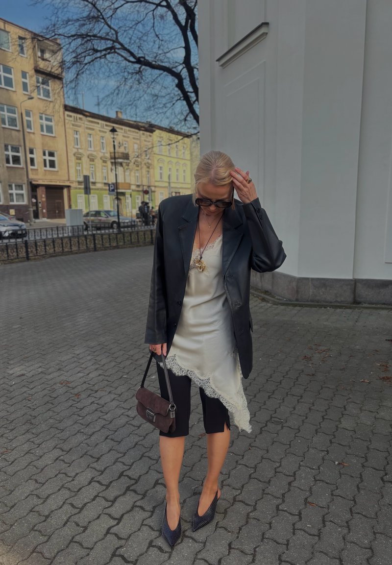 Femeie care își aranjează părul, purtând un blazer negru supradimensionat, o rochie albă de tip slip cu dantelă, pantaloni capri negri, pantofi cu toc negri și ținând o geantă de mână închisă la culoare, în aer liber.
