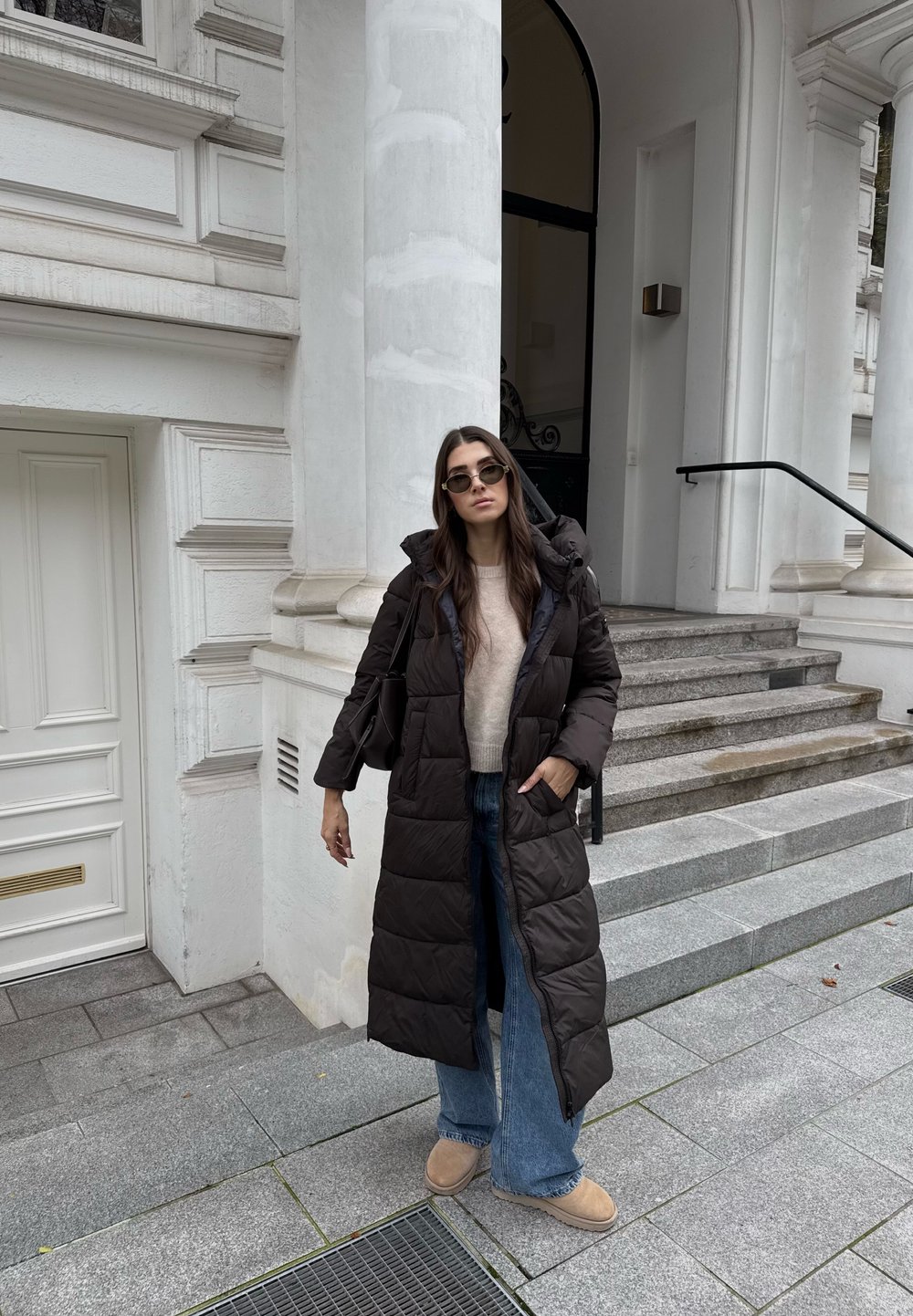 Tumed pruun puffer-kate koos kapuutsiga, kombineeritud heleda kootud kampsuni ja laia jalgadega siniste teksadega. Taustal on valge arhitektuur ja astmed.