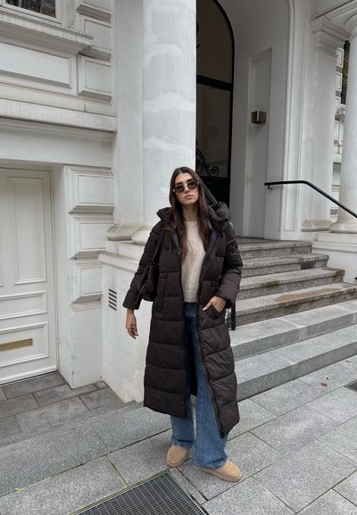 Abrigo acolchado marrón oscuro con capucha, combinado con un suéter ligero y jeans de pierna ancha azules. El fondo presenta arquitectura blanca y escaleras.