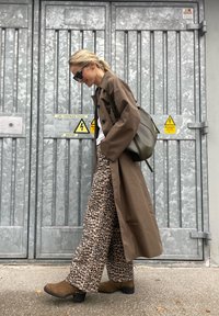 Brown long trench coat, leopard print wide-leg trousers, brown ankle boots. Black sunglasses, olive green bag, grey metal background.