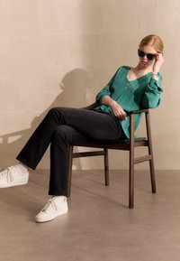Grüne Bluse mit Knöpfen, schwarze Jeans und weiße Sneakers. Das Modell trägt Sonnenbrillen und sitzt lässig auf einem Holzstuhl. Neutrale Hintergrund.