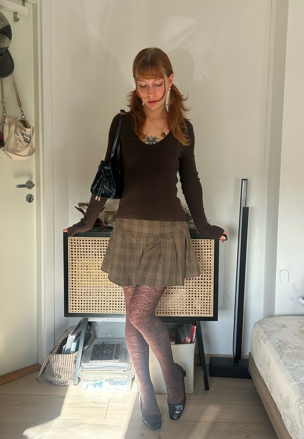 Une femme pose à côté d’un meuble à façade en cannage. Elle porte un haut foncé, une jupe à carreaux, des collants à motifs et des chaussures à talons.