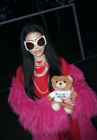 Red dress, pink fur coat, oversized white sunglasses. Pearls layered around the neck. Holding a brown teddy bear in a white shirt with text.