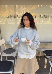 Sivi prevelik pulover z zapletenimi belimi čipkastimi manšetami, nošen preko bele srajce z gumbi in bež krila, drži belo skodelico kave.