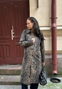 Kabát s leopardím vzorom z mäkkej látky, s golierom a gombíkmi. Kombinovaný s tmavými nohavicami a čiernou kabelkou. Betonové pozadie.