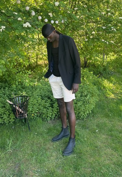 Blazer negro sobre una camiseta sin mangas negra, pantalones cortos beige claros y botas negras hasta el tobillo. De pie junto a una cesta negra en un entorno natural verde.