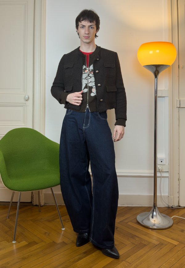 Ung person stående indendørs iført en sort cropped jakke, brede mørke jeans og støvler, med en grøn stol og en gulvlampe i nærheden.