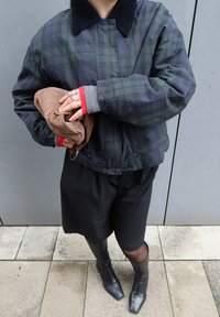 Personne portant une veste à carreaux foncés, une jupe noire et des bottes, tenant un sac plié marron devant un mur gris.