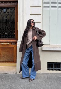 Pruun ülisuur mantel kreemja kampsuni kohal, kombineeritud laiade siniste teksade ja pruunide teravate saabastega, aksessuaarina põimitud kott.