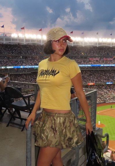 Camisa amarilla de cuello redondo con texto gráfico negro, falda de camuflaje, gorra beige y bolso negro; sentado en un estadio con espectadores.